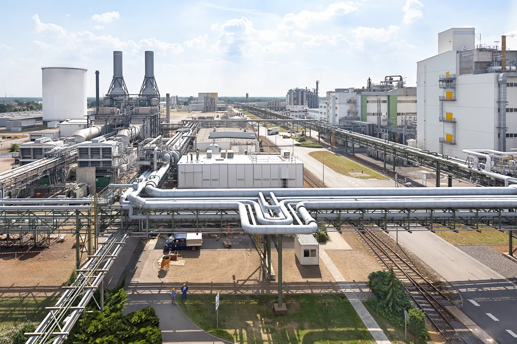 A large industrial plant seen from above: pipe racks, silos, process buildings and rail sidings under a bright sky.